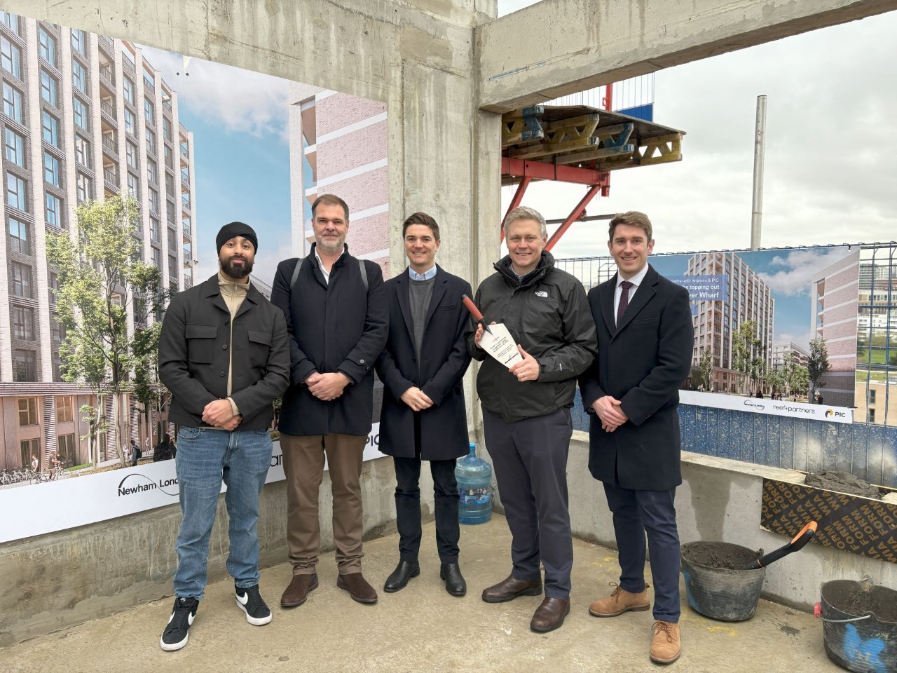 five men standing for photo, one holding builders trowel on construction site.